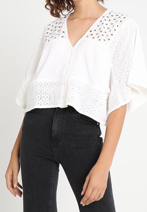 Žena v bílé blůze z dírkované krajky se zapínáním na knoflíky a širokými rukávy, doplněné o černé džíny s vysokým pasem, na jednobarevném pozadí.