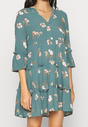 Robe florale bleu sarcelle avec volants à plusieurs niveaux et manches trois-quarts, portée par une personne, montrant le buste et les bras sur un fond uni.
