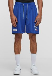 Blå mesh basketballshorts med snøre i livet, med "NYC" grafik og et logo patch. Båret med hvite sokker og sneakers.