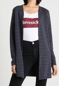 Gilet rayé bleu marine à ouverture devant, à manches longues et en tissu texturé, porté sur une chemise blanche avec des lettres rouges, associé à un pantalon noir.