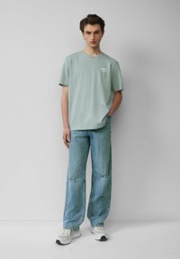 Jongeman in een lichtgroene grafische t-shirt, losse gescheurde blauwe jeans en witte sneakers, staand tegen een effen lichtgrijze achtergrond.