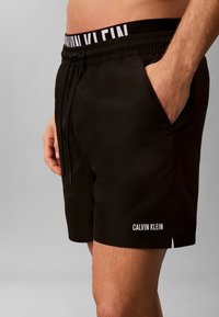 Мъжки торс и горни крака, носещи черни шорти Calvin Klein с джобове отстрани и видима лента с марка под гола кожа.