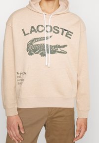 Felpa con cappuccio beige con tasca frontale, con grafica di un coccodrillo verde scuro e scritta "LACOSTE". Coulisse bianche e polsini a costine.