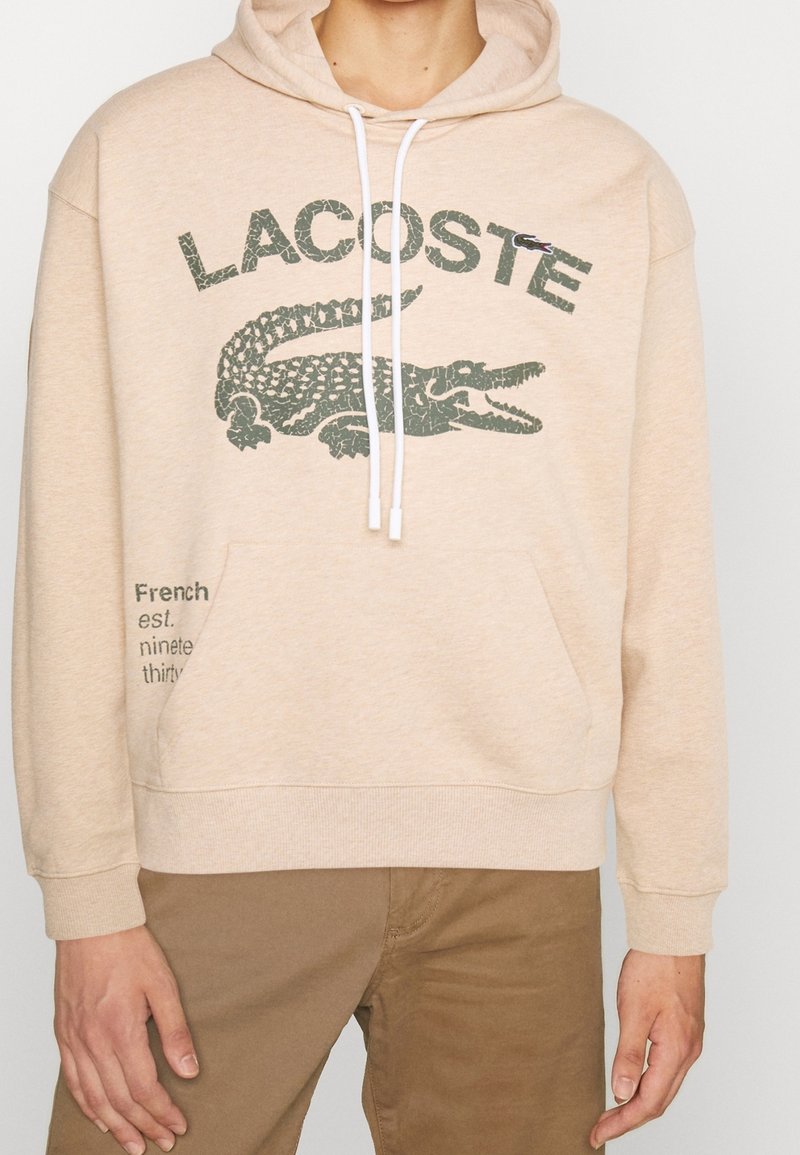 Felpa con cappuccio beige con tasca frontale, con grafica di un coccodrillo verde scuro e scritta "LACOSTE". Coulisse bianche e polsini a costine.