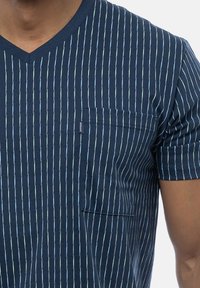 Mand iført navyblå V-hals T-shirt med fine lodrette lyseblå og grønne striber og en forlommeslomme på højre side.