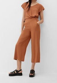 Ensemble deux pièces couleur rouille avec un haut à manches courtes et un pantalon à jambes larges, fabriqué en tissu lisse ; associé à des sandales noires épaisses.