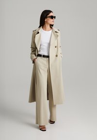 Beige double-breasted trench coat with gold buttons, worn over a white shirt. Paired with wide-leg beige trousers and black sandals.