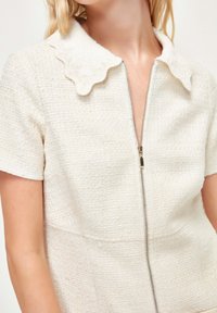Vestido blanco de manga corta con un tejido texturizado, un cuello festoneado y un cierre de cremallera en la parte delantera. Los detalles incluyen un delicado bordado.