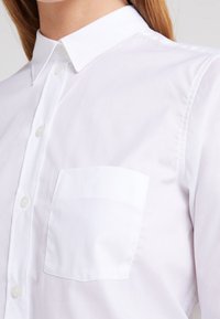 Camicia bianca a maniche lunghe con colletto classico, taschino sul petto e texture liscia. Presenta bottoni rotondi e vestibilità sartoriale.