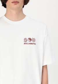 Balta medvilninė marškinėliai su priekinio logo, kuriame pavaizduotas medis, yin-yang ir saulės ikonų, visi vyšninių spalvos, su tekstu "BILLABONG" apačioje.