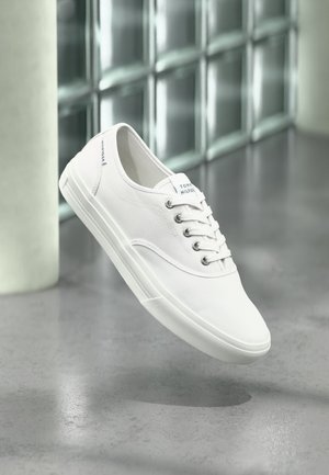 Baskets basses blanches en toile avec lacets, étiquetées Tommy Hilfiger, montrées flottant au-dessus d’un sol gris lisse près d’un mur en briques de verre.