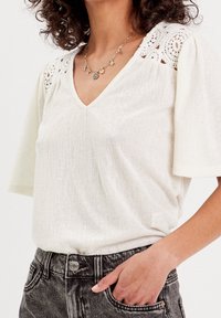 Haut blanc à manches courtes avec un col en V, agrémenté d'accents en dentelle sur les épaules et d'un tissu texturé. Associé à un jean foncé taille haute.