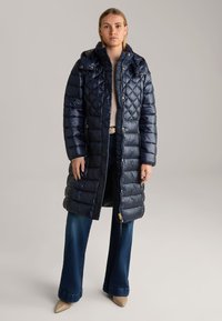 Marineblauwe gewatteerde jas met een quiltpatroon, hoge kraag, gouden hardware accenten en een lang ontwerp, gecombineerd met flared jeans en hakken.