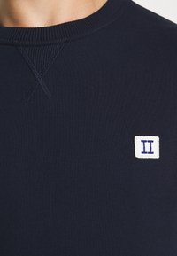 Maglione girocollo blu navy con dettagli di cucitura visibili e una piccola toppa bianca con due linee verticali blu sul petto.