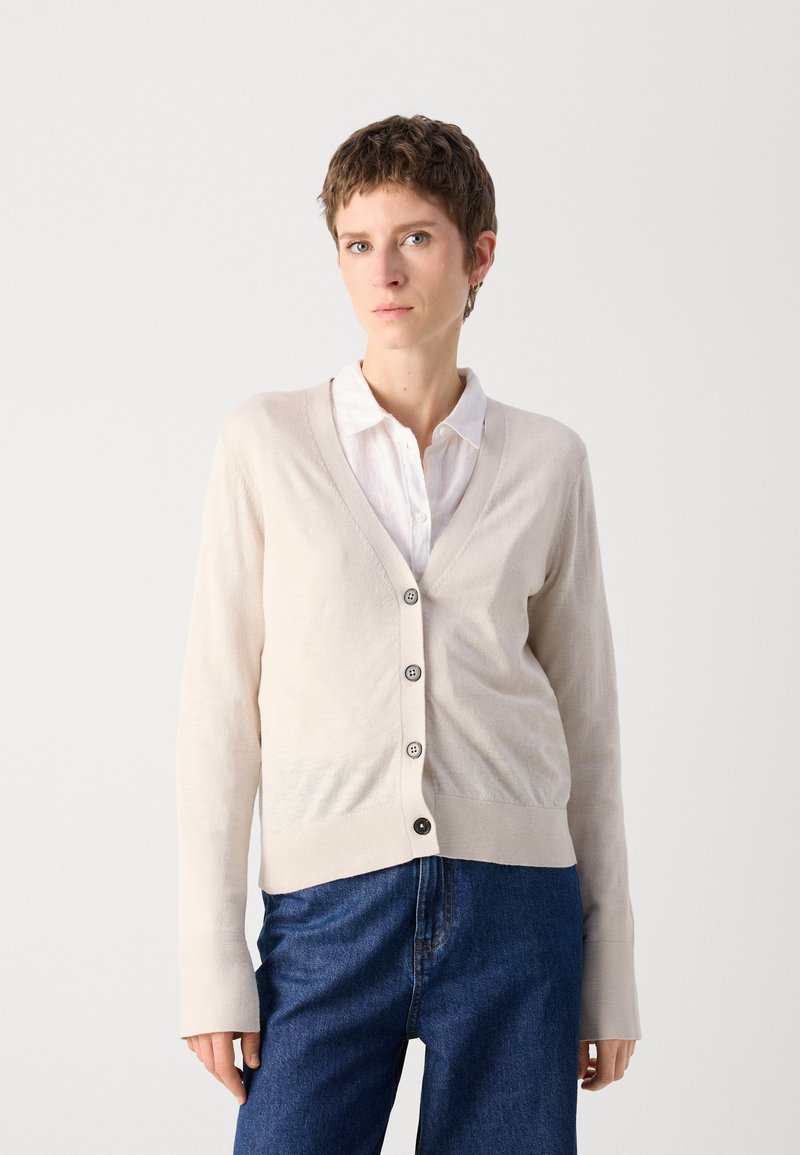 Beige Strickjacke aus weichem Material, mit V-Ausschnitt, langen Ärmeln und fünf schwarzen Knöpfen auf der Vorderseite, kombiniert mit blauen Jeans.