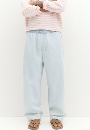 Osoba nosząca jasnoniebieskie, luźne dżinsy i beżowe sandały, z założonymi na piersi rękami na różowo-białej paskowanej koszulce z długim rękawem.