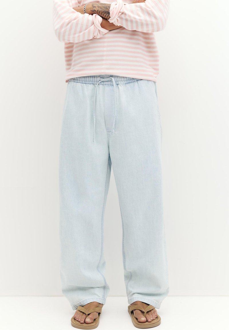 Personne portant un jean bleu clair ample et des sandales beige, les bras croisés sur une chemise à manches longues rayée rose et blanche.