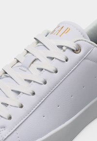 Λευκό δερμάτινο αθλητικό παπούτσι με λευκά κορδόνια, μικρές τρύπες στο πλάι και λογότυπο «GAP» σε χρυσό στο γλώσσα.