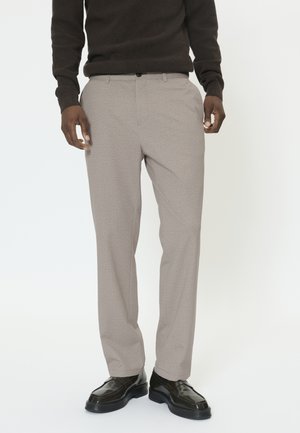 Pantalon beige en tissu texturé. Il présente une coupe droite, des poches avant et une fermeture à bouton. Associé à des chaussures foncées et brillantes.