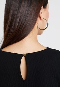 Top nero texturizzato con scollatura rotonda, caratterizzato da un piccolo apertura a cuore sul retro chiuso da un accento in bottone dorato.