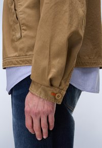 Καφέ, ανθεκτικό μπουφάν με κουμπωτή μανσέτα, πορτοκαλί ραφές και άνετη εφαρμογή, φορεμένο πάνω από ένα ανοιχτό μπλε πουκάμισο με γιακά και σκούρο τζιν.