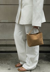 Borsa a mano in suede color sabbia con forma strutturata e manici corti, abbinata a un completo bianco oversize e sandali piatti beige chiaro.
