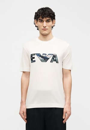 Fiatal férfi rövid, göndör hajjal, fehér, rövid ujjú pólót visel, amelyen nagy "EVA" felirat látható kék terepmintával.