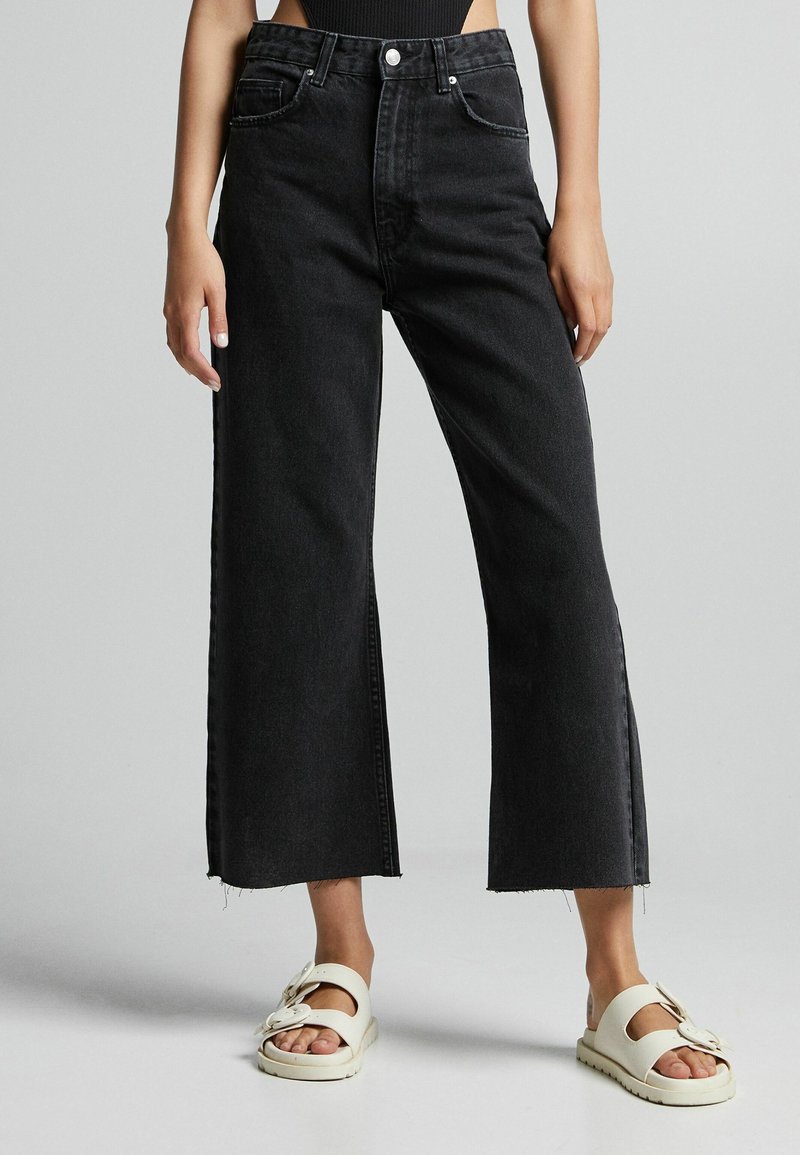 Svart, högmidjade vida jeans med rå nederkant och sidofickor, tillverkade av denimmaterial; kombinerat med vita slip-on-sandaler.