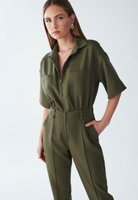 Olijfgroene jumpsuit met korte mouwen, voorkeursrits, twee borstzakken en een elastische tailleband. Gemaakt van een gladde stof.