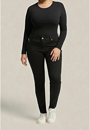 Vrouw draagt een nauwsluitende zwarte top met lange mouwen, een zwarte broek met hoge taille en witte sneakers tegen een effen achtergrond.