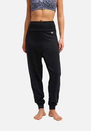 Femme portant un pantalon de jogging noir taille haute et un soutien-gorge de sport à motifs, debout pieds nus sur un fond uni.
