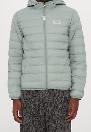 Mann trägt eine hellgraue, gesteppte Jacke mit Reißverschluss und Kapuze sowie gemusterte dunkle Hosen mit wiederholten "EA7"-Logos.