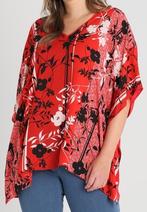 Rød løs bluse med sort-hvide blomsterprint, båret med blå jeans, viser overkroppen og armen.