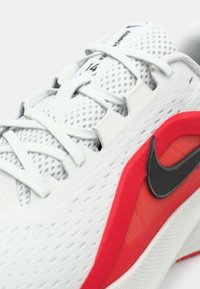 Chaussure de sport en mesh blanc avec des accents rouges et le swoosh Nike noir, avec des lacets blancs et une étiquette de taille 14 sur la languette.