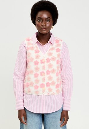 Donna con afro che indossa una camicia a righe rosa con bottoni sotto un gilet bianco con motivi floreali rosa in rilievo, abbinata a jeans azzurri.