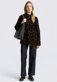 Pull imprimé léopard marron en tissu texturé, associé à un jean en denim foncé et des bottines noires, tenant un sac à main noir souple.