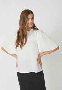 Blusa bianca con maniche corte e svasate, dettagli plissettati attorno al collo e un tessuto testurizzato. Indossata con una gonna nera.