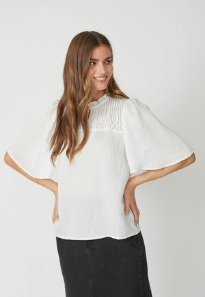 Blusa bianca con maniche corte e svasate, dettagli plissettati attorno al collo e un tessuto testurizzato. Indossata con una gonna nera.