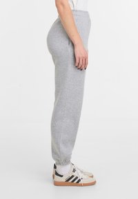 Pantalons de survêtement gris avec une jambe fuselée, une taille élastique et des poignets. Portés avec des baskets légères à rayures noires et une base beige.