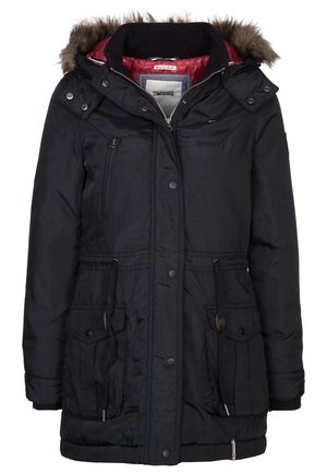Femme portant un parka noir à capuche avec bordure en fourrure, un pull en tricot blanc, un jean bleu et des bottines marron à lacets, debout sur un fond uni.