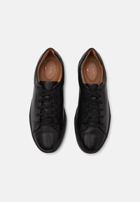 Adidași din piele neagră cu o textură netedă, vârf rotund și șireturi negre. Au o branț bej cu logo-ul brandului. Design cu profil scăzut.