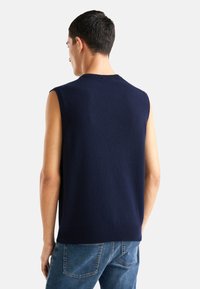Granatowy bezrękawnik z dzianiny o okrągłym dekolcie, miękkiej fakturze i ściśniętym wykończeniu. Noszony na jasnoniebieskich dżinsach widok od tyłu.