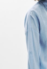 Camicia a maniche lunghe di lino di colore azzurro chiaro. Presenta un polsino con bottoni e un tessuto liscio e strutturato con un sottile motivo a tessitura.