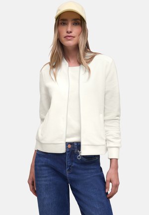 Kvinde iført en cremefarvet trøje med lynlås, hvid skjorte, blå jeans og en lysebeige baseballkasket, der står foran en ensfarvet baggrund.