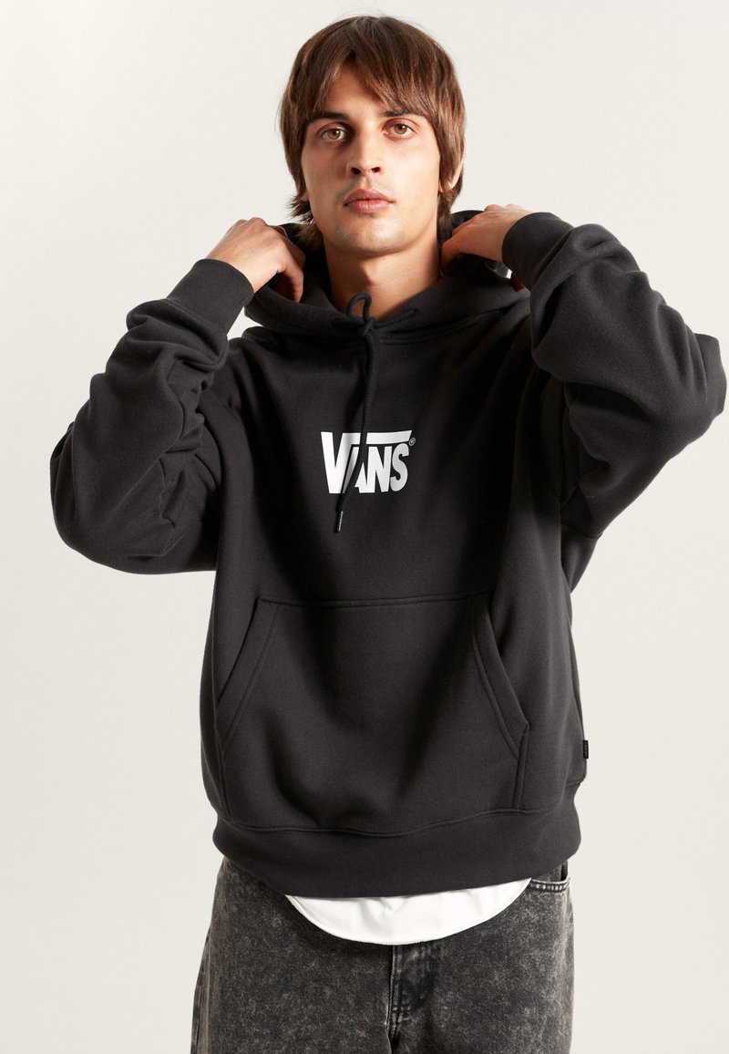 Sudadera negra de Vans con un bolsillo frontal, capucha ajustable con cordón y logo blanco. Tejido suave con un corte relajado, llevada sobre una camiseta blanca.