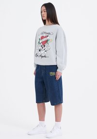 Ljusgrå sweatshirt med dödskalle- och hjärtmotiv, ihopparad med lösa denimshorts som har gul text. Vita sneakers fulländar outfiten.