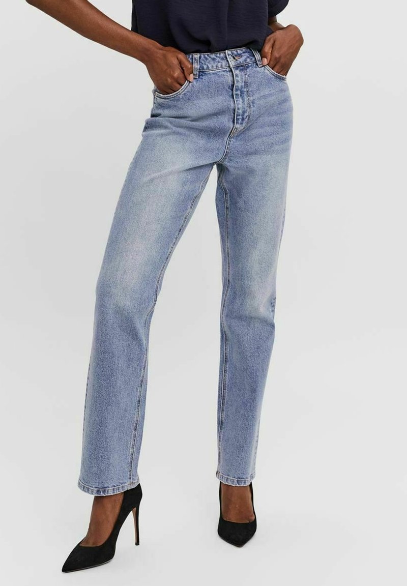 Femme portant un jean bleu clair taille haute à coupe droite, les mains dans les poches et des talons hauts noirs pointus sur fond blanc.