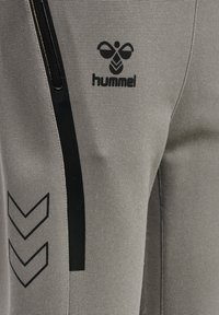 Graue Sporthose aus strukturiertem Material, mit einer schwarzen Reißverschlusstasche, schwarzem Logo und schwarzen Chevron-Akzenten an der Seite.