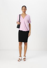 Top rose clair à col en V, jupe noire cintrée et sandales à talons carrés blanches. Le modèle tient un sac à main noir, se tenant devant un fond blanc.