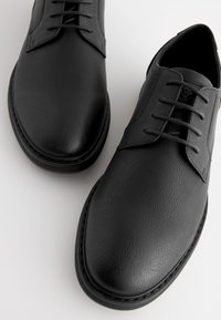 Chaussures à lacets en cuir noir avec une finition texturée, bout arrondi et détails surpiqués. Design minimaliste avec une silhouette lisse et épurée.